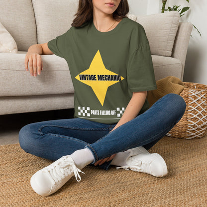 A woman sitting cross-legged on a woven rug in front of a beige sofa, wearing an green graphic T-shirt with a large yellow star and the text 'VINTAGE MECHANIC' and 'PARTS FALLING OFF'.