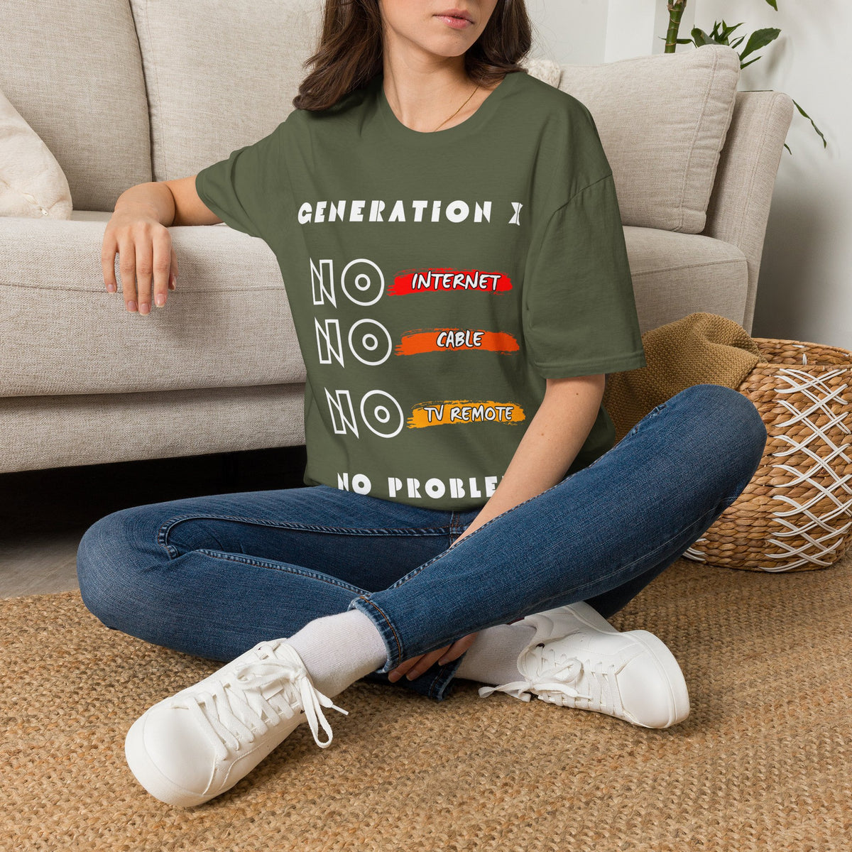 A woman sitting cross-legged on a woven rug in front of a beige sofa, wearing an green graphic T-shirt with white text "GENERATION X NO INTERNET, NO CABLE, NO TV REMOTE NO PROBLEM" paired with blue jeans and white sneakers.
