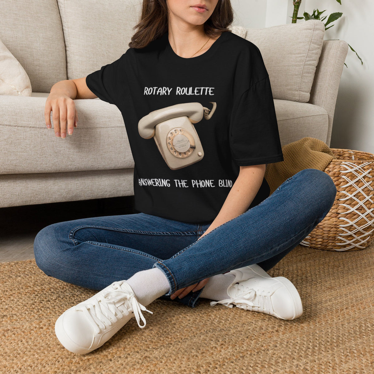 A woman sat on a rug next to a couch wearing a black graphic t-shirt featuring a black rotary telephone and the text 'ROTARY ROULETTE' and 'ANSWERING THE PHONE BLIND'. 