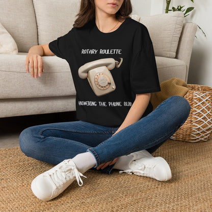A woman sat on a rug next to a couch wearing a black graphic t-shirt featuring a black rotary telephone and the text 'ROTARY ROULETTE' and 'ANSWERING THE PHONE BLIND'. 