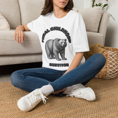 A woman sitting on a woven rug in front of a beige sofa, wearing a white graphic t-shirt with a black bear illustration and the text 'FERAL CHILDHOOD SURVIVOR', paired with blue jeans and white sneakers.