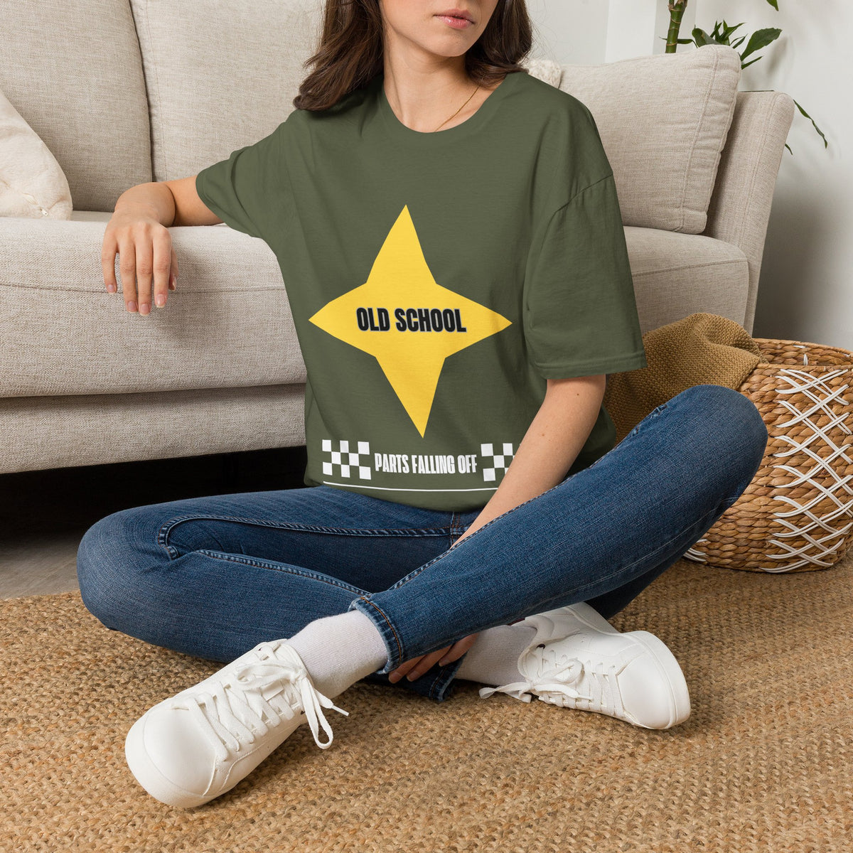 A woman sitting cross-legged on a woven rug in front of a beige sofa, wearing a green graphic T-shirt with a large yellow star and the text 'OLD SCHOOL' and 'PARTS FALLING OFF'.