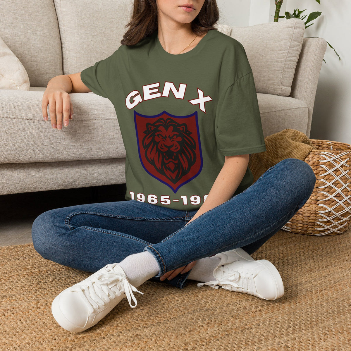 A woman sitting cross-legged on a woven rug in front of a beige sofa, wearing a green graphic T-shirt with a white 'GEN X' text and a red-and-purple shield emblem, paired with blue jeans and white sneakers.
