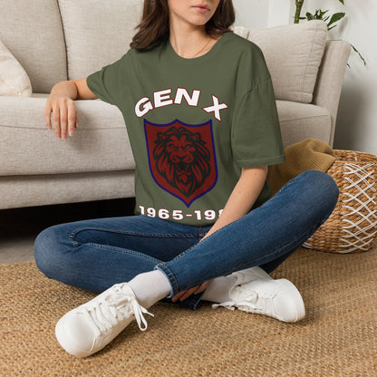 A woman sitting cross-legged on a woven rug in front of a beige sofa, wearing a green graphic T-shirt with a white 'GEN X' text and a red-and-purple shield emblem, paired with blue jeans and white sneakers.