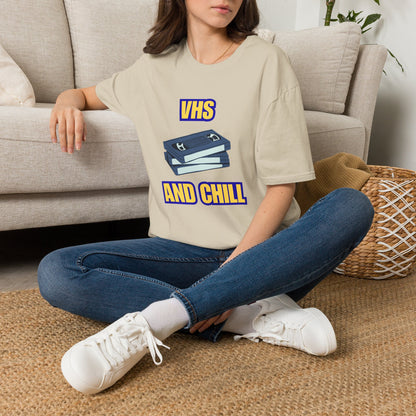 A woman sitting cross-legged on a woven rug in front of a beige sofa, wearing a sand graphic T-shirt with a blue and yellow VHS stack design and the words 'VHS AND CHILL'.