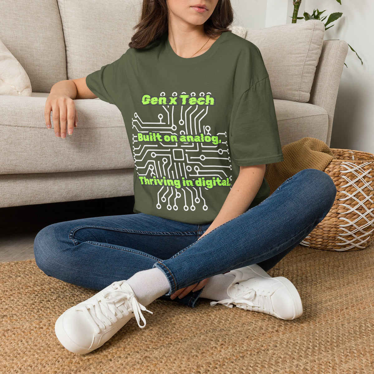 A woman sits cross-legged on a woven rug in a living room, wearing a green graphic T-shirt with a white circuit board design and the green text 'Gen X Tech Built on analog Thriving in digital.' She is also wearing blue jeans and white sneakers.
