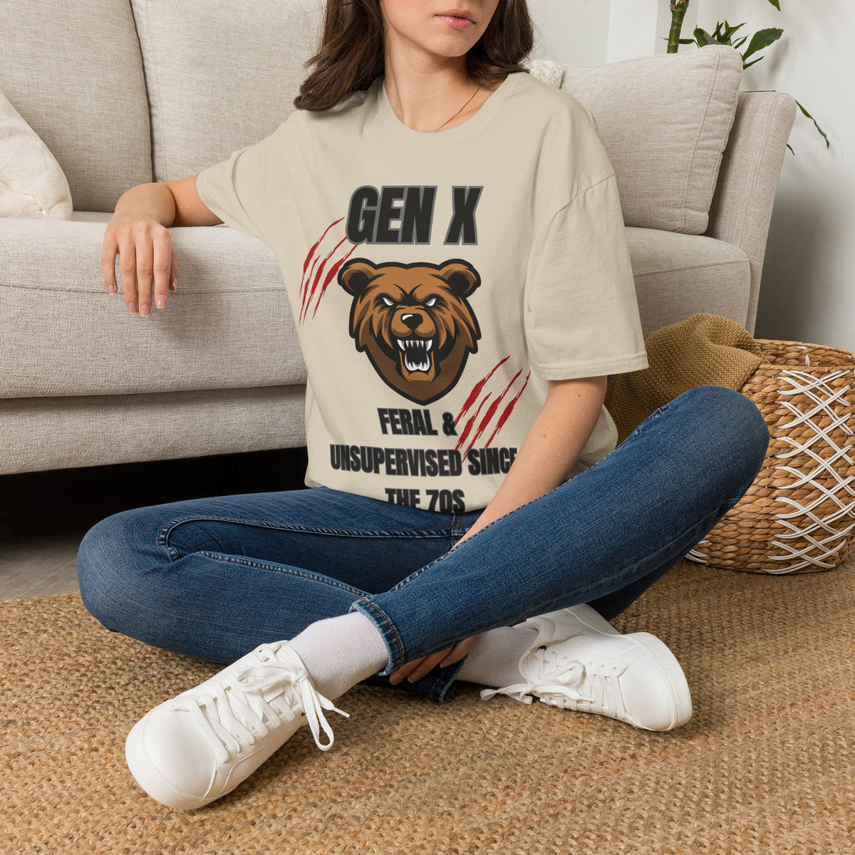 A woman sitting cross-legged on a woven rug in front of a light beige sofa, wearing a sand graphic t-shirt with a brown bear design and the text 'GEN X FERAL & UNSUPERVISED SING THE 7OS'.