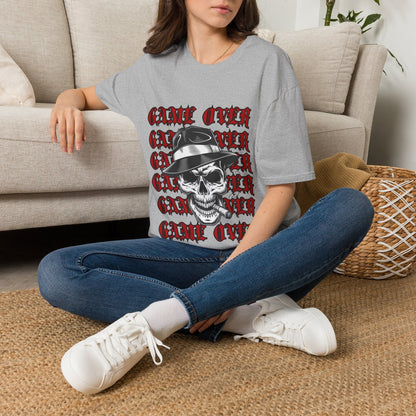 A woman sitting cross-legged on a woven rug in front of a beige sofa, wearing a gray oversized graphic T-shirt with a red and black skull design and the words 'GAME OVER' repeated in red gothic lettering.