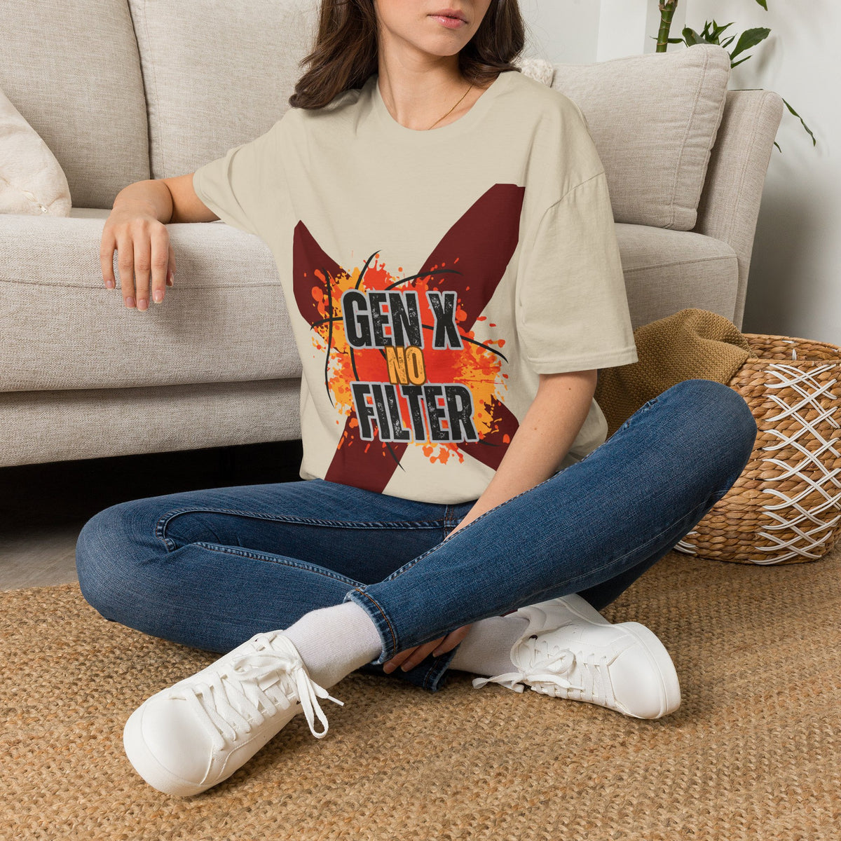 A woman sits cross-legged on a woven rug in front of a light beige sofa, wearing an oversized sand graphic T-shirt with blue jeans and white sneakers. The shirt features a large red 'X' with orange paint-splatter details and the text 'GEN X NO FILTER'