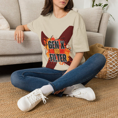 A woman sits cross-legged on a woven rug in front of a light beige sofa, wearing an oversized sand graphic T-shirt with blue jeans and white sneakers. The shirt features a large red 'X' with orange paint-splatter details and the text 'GEN X NO FILTER'