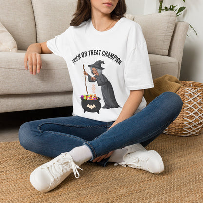 A woman sitting on a woven rug in front of a beige couch, wearing a white graphic T-shirt with a witch and cauldron design and the text 'TRICK OR TREAT CHAMPION', paired with blue jeans and white sneakers.