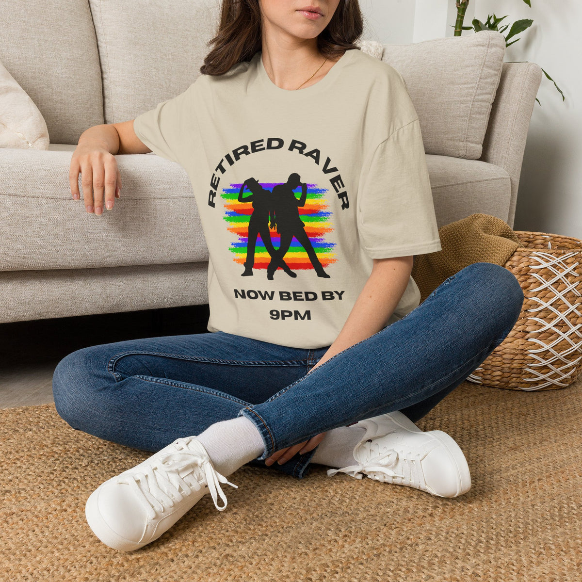 A woman sitting cross-legged on a woven rug in front of a beige sofa, wearing a sand graphic T-shirt with the text 'RETIRED RAVER NOW BED BY 9PM' and a black silhouette over a rainbow design, paired with blue jeans and white sneakers.