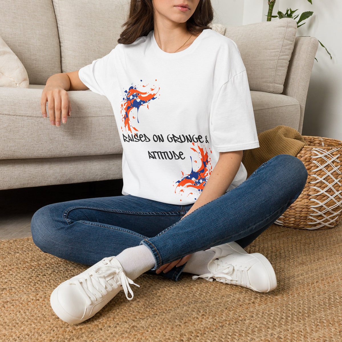 A woman sitting cross-legged on a woven rug in front of a beige sofa, wearing a white graphic T-shirt with blue jeans and white sneakers. The T-shirt features a red and blue paint splatter design with the text 'RAISED ON GRUNGE & ATTITUDE'.
