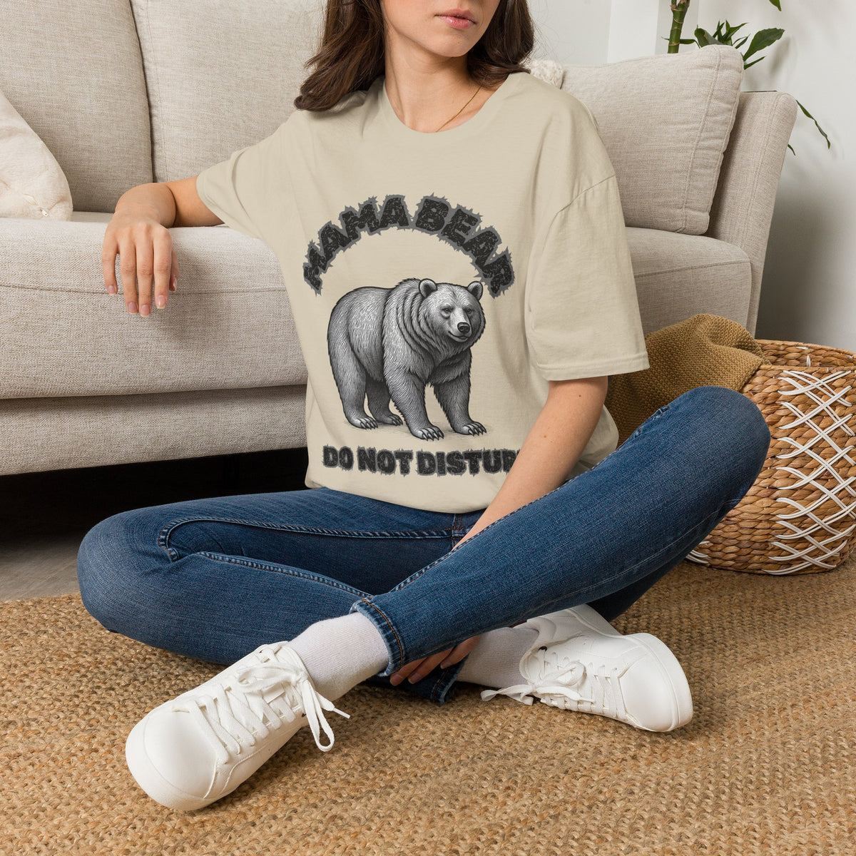 A woman sitting cross-legged on a woven rug in front of a light beige sofa, wearing a sand graphic t-shirt with a black bear print and the text 'MAMA BEAR DO NOT DISTURB', paired with blue jeans and white sneakers.