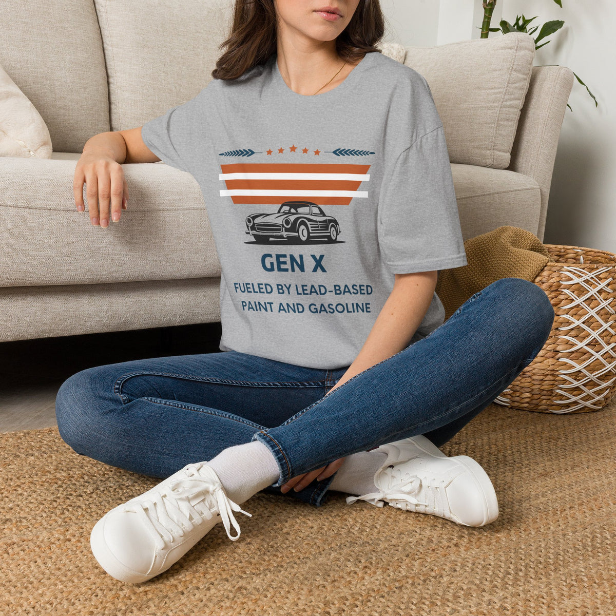 A woman sat on rug next to couch wearing a grey graphic t-shirt. The t-shirt features a retro design with the text 'GEN X FUELED BY LEAD-BASED PAINT AND GASOLINE' and an illustration of a classic car under orange and white stripes.