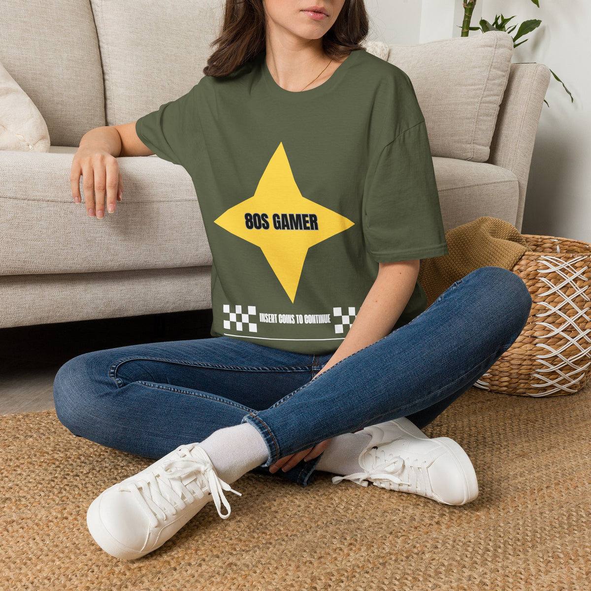 A woman sitting cross-legged on a woven rug in front of a beige sofa, wearing a green graphic T-shirt with a large yellow star and the text '80S GAMER in black, and a white text bar below reading 'INSERT COINS TO CONTINUE' flanked by black-and-white checkered squares.