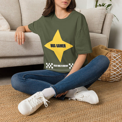 A woman sitting cross-legged on a woven rug in front of a beige sofa, wearing a green graphic T-shirt with a large yellow star and the text '80S GAMER in black, and a white text bar below reading 'INSERT COINS TO CONTINUE' flanked by black-and-white checkered squares.