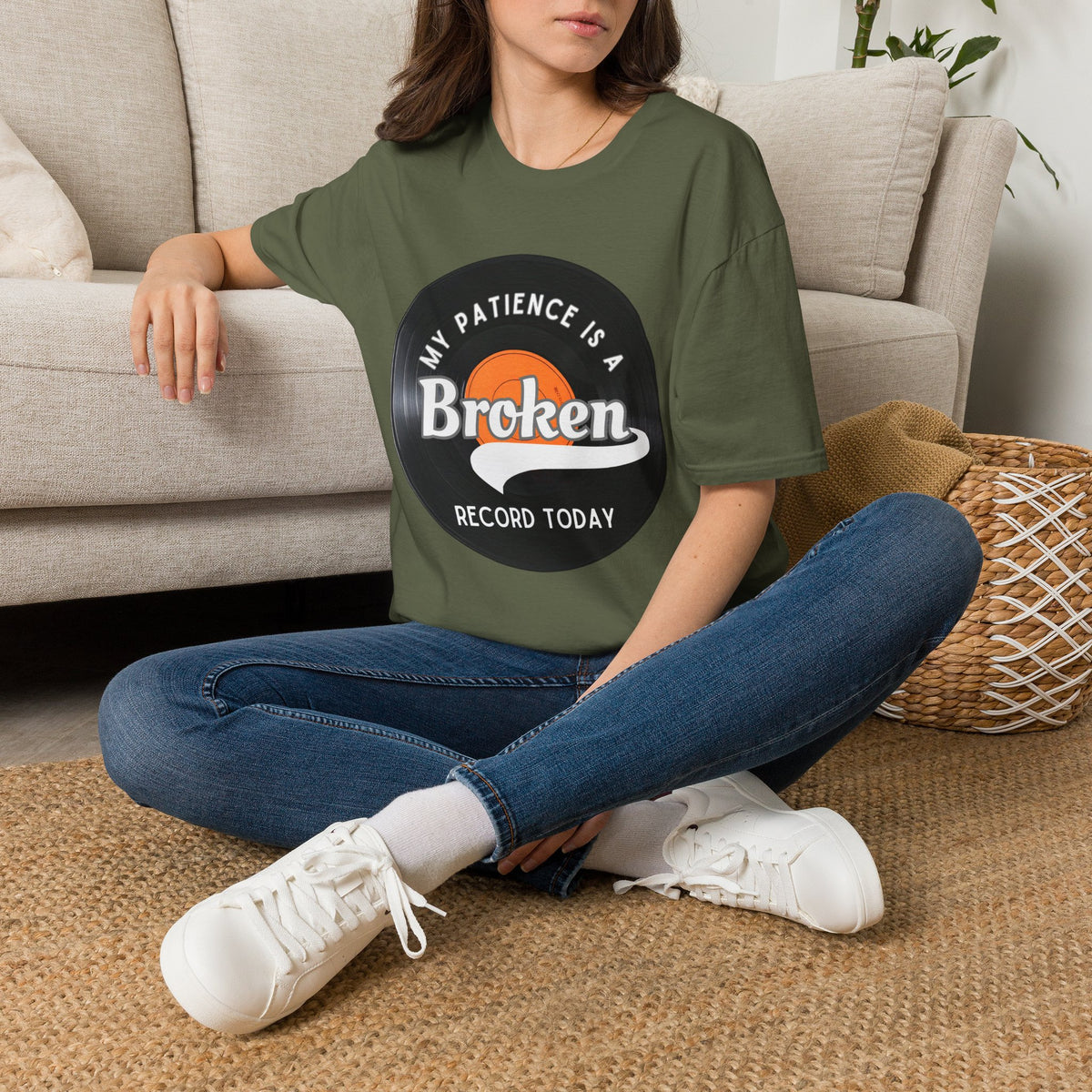 A woman sitting cross-legged on a woven rug in front of a beige sofa, wearing an green T-shirt with a vinyl record graphic and text "My pateince is a broken record today"