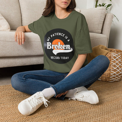 A woman sitting cross-legged on a woven rug in front of a beige sofa, wearing an green T-shirt with a vinyl record graphic and text "My pateince is a broken record today"