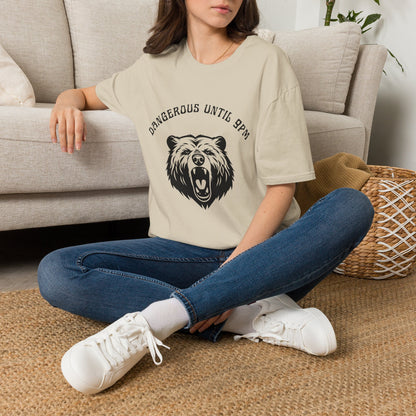 A woman sitting cross-legged on a woven rug in front of a beige sofa, wearing a sand graphic T-shirt with a black bear head design that reads 'Dangerous till 9pm'