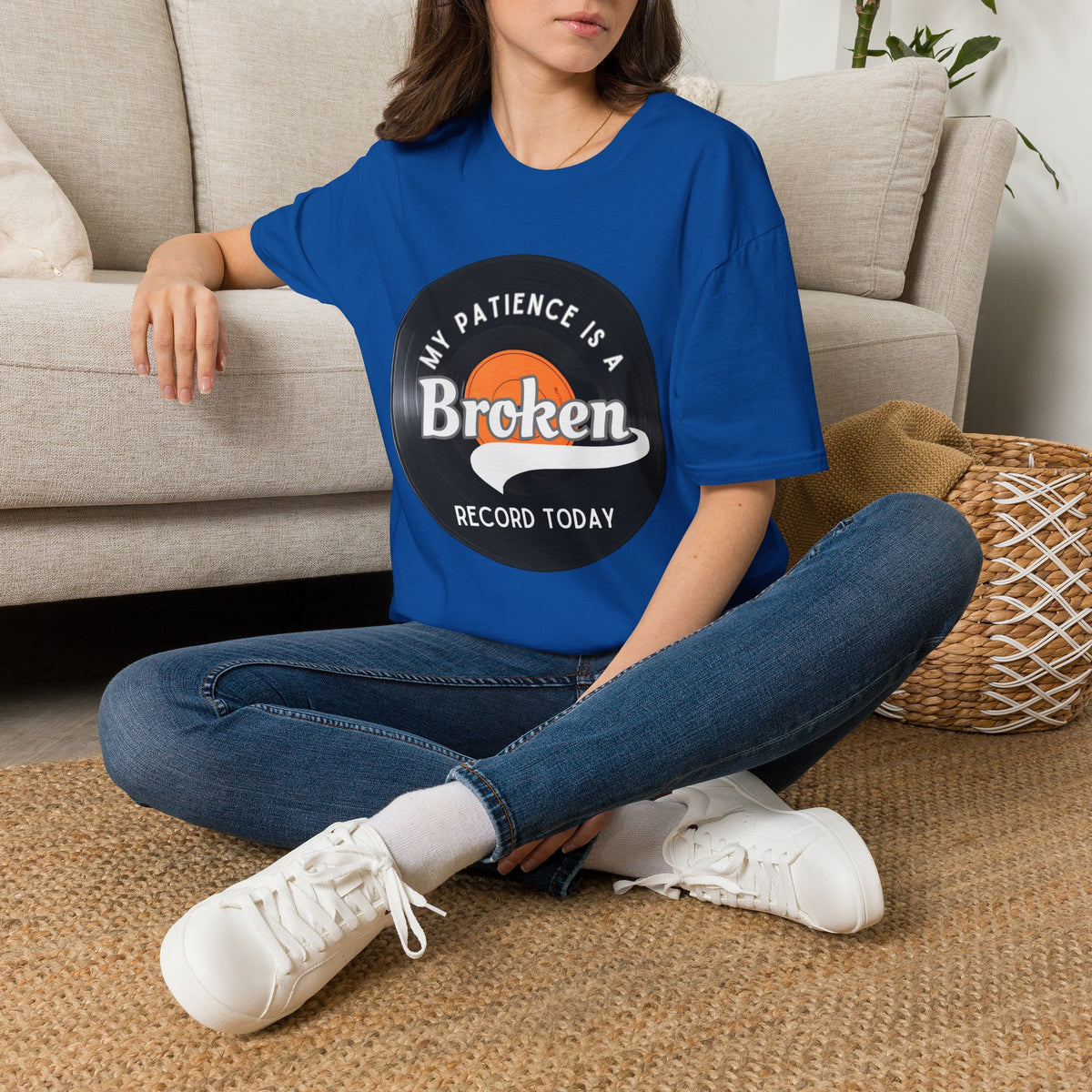 A woman sitting cross-legged on a woven rug in front of a beige sofa, wearing a royal blue graphic T-shirt with a vinyl record graphic and text that reads 'MY PATIENCE IS A BROKEN RECORD TODAY.'.