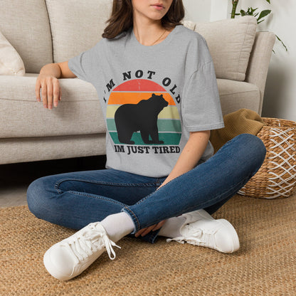 A woman sitting cross-legged on a woven rug in front of a beige sofa, wearing a gray graphic T-shirt with a bear silhouette and the text 'I'M NOT OLD I'M JUST TIRED' over a retro sunset design, paired with blue jeans and white sneakers.