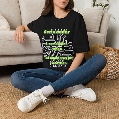 A woman sitting cross-legged on a woven rug in front of a beige sofa, wearing a black graphic T-shirt with green text and circuit board artwork reading 'Gen X coder I remember when the cloud was just weather.' She is also wearing blue jeans and white sneakers.