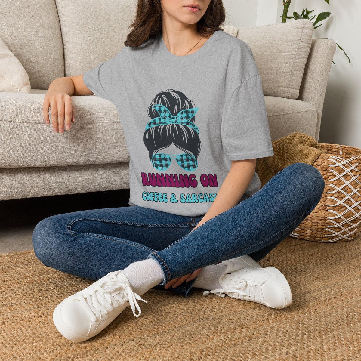 A woman sitting on a woven rug in front of a beige sofa, wearing a gray graphic t-shirt with a cartoon hair bun and the text 'RUNNING ON COFFEE & SALTARY'.