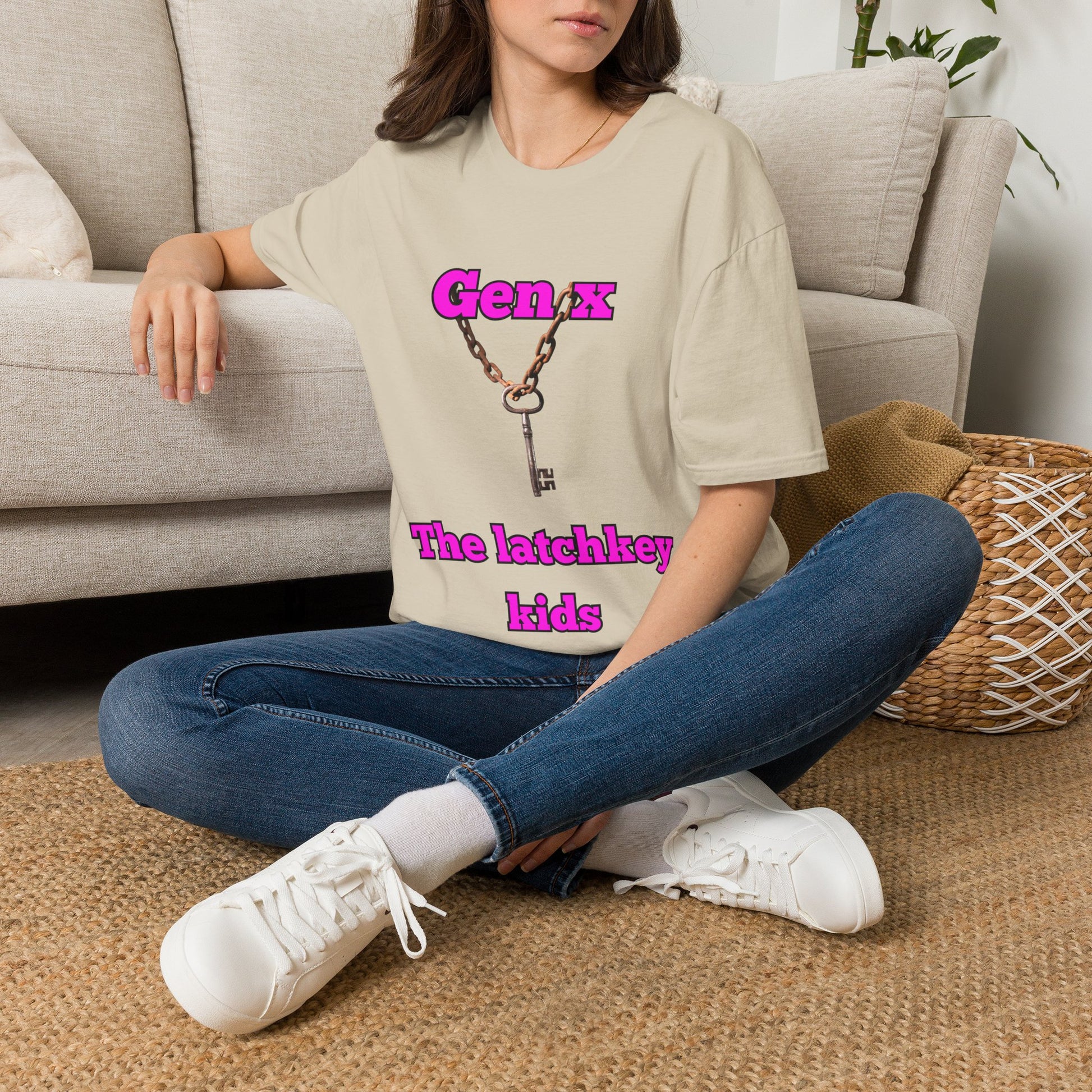 A woman sitting cross-legged on a woven rug in front of a light gray sofa, wearing an oversized sand graphic T-shirt with pink text reading 'Gen x The latchkey kids' and blue jeans with white sneakers.