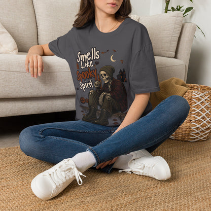 A woman sitting cross-legged on a woven rug in front of a beige sofa, wearing a dark gray graphic T-shirt with a skeleton and the text 'Smells Like Spooky Spirit.'.