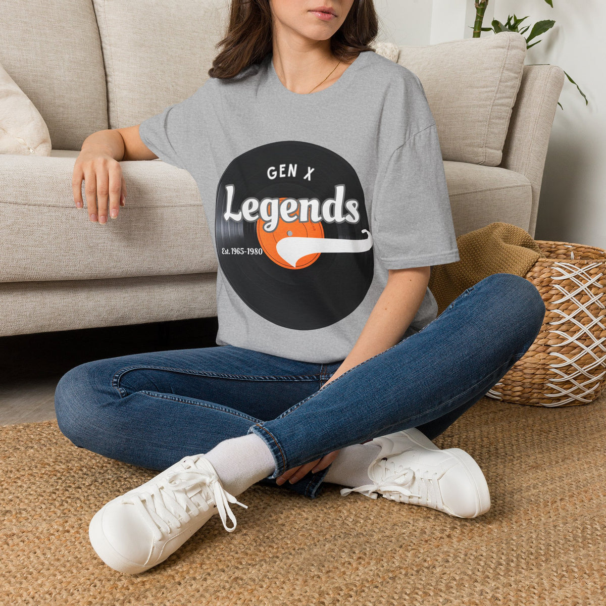 A woman sitting cross-legged on a woven rug in front of a beige sofa, wearing a gray graphic T-shirt with a vinyl record that reads 'GEN X Legends' 