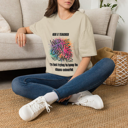 A woman sits cross-legged on a woven rug in front of a light beige sofa, wearing an oversized sand graphic T-shirt with colorful graphic and "GEN X TEACHER IM JUST TRYING TO KEEP THE CHAOS COLOURFUL" blue jeans with white sneakers.