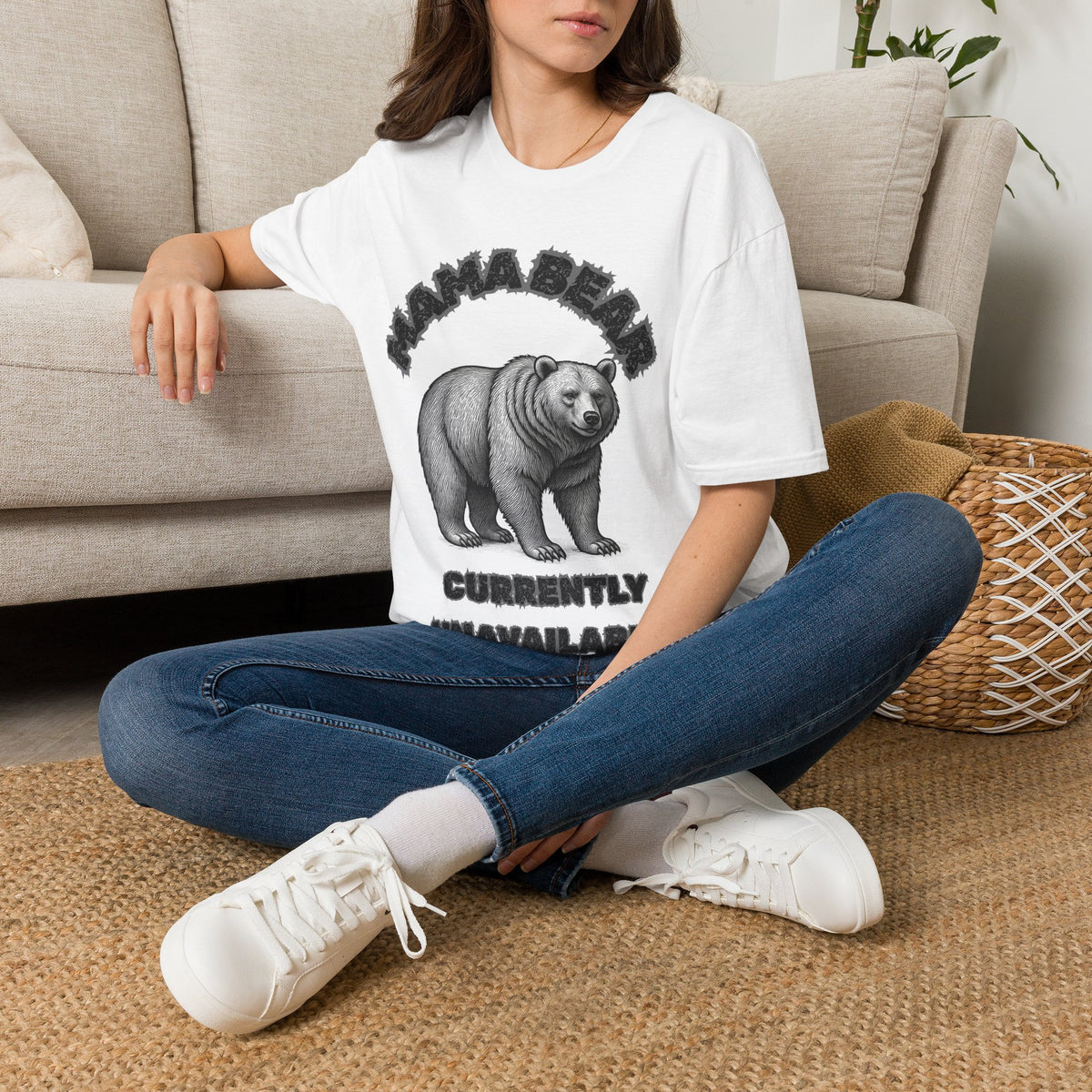 A woman sitting cross-legged on a woven rug in front of a beige sofa, wearing an oversized white graphic T-shirt with a black bear print and the text 'MAMA BEAR CURRENTLY UNAVAILABLE', paired with blue jeans and white sneakers.