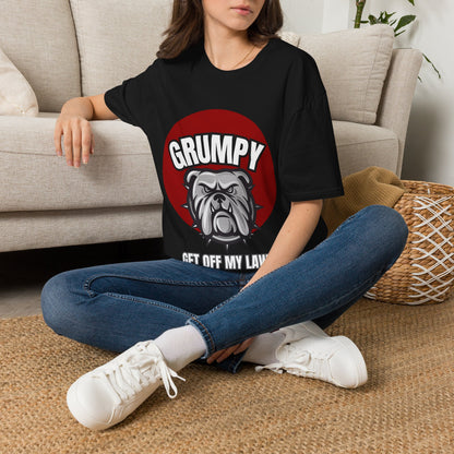 A woman sitting cross-legged on a woven rug in front of a beige sofa, wearing a black graphic T-shirt with a red circle, white text reading 'GRUMPY GET OFF MY LAW,' and a bulldog illustration. She is also wearing blue jeans and white sneakers.