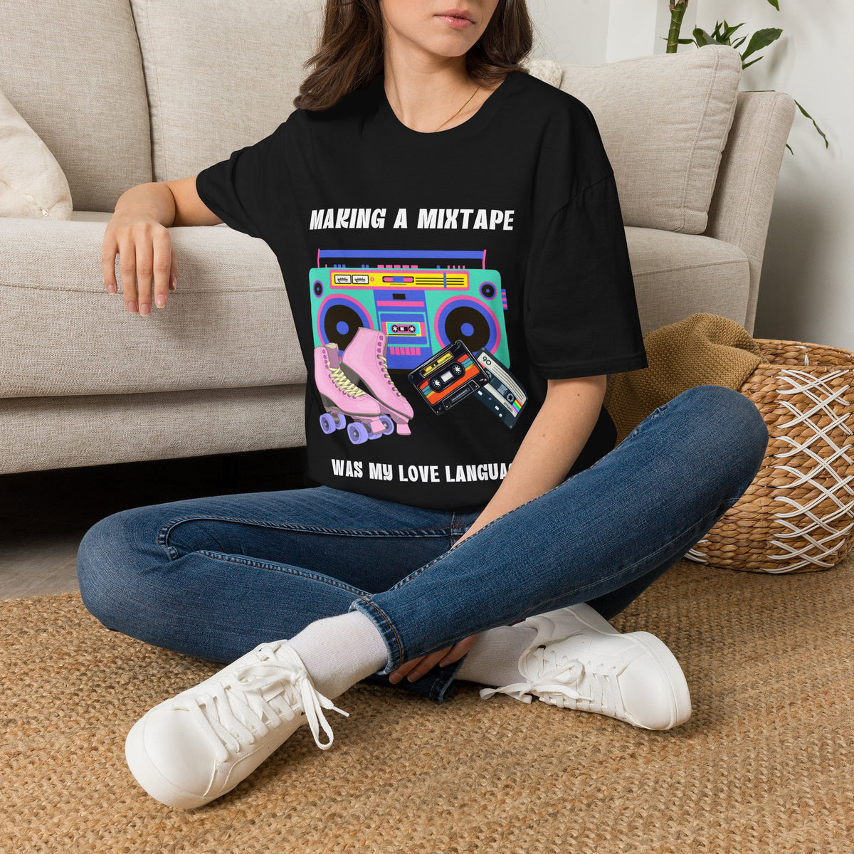 A woman sitting on a woven rug in front of a beige sofa, wearing a black graphic t-shirt with a colorful retro boombox, cassette tape, roller skate, and text that reads 'MAKING A MIXTAPE WAS MY LOVE LANGUAGE'.