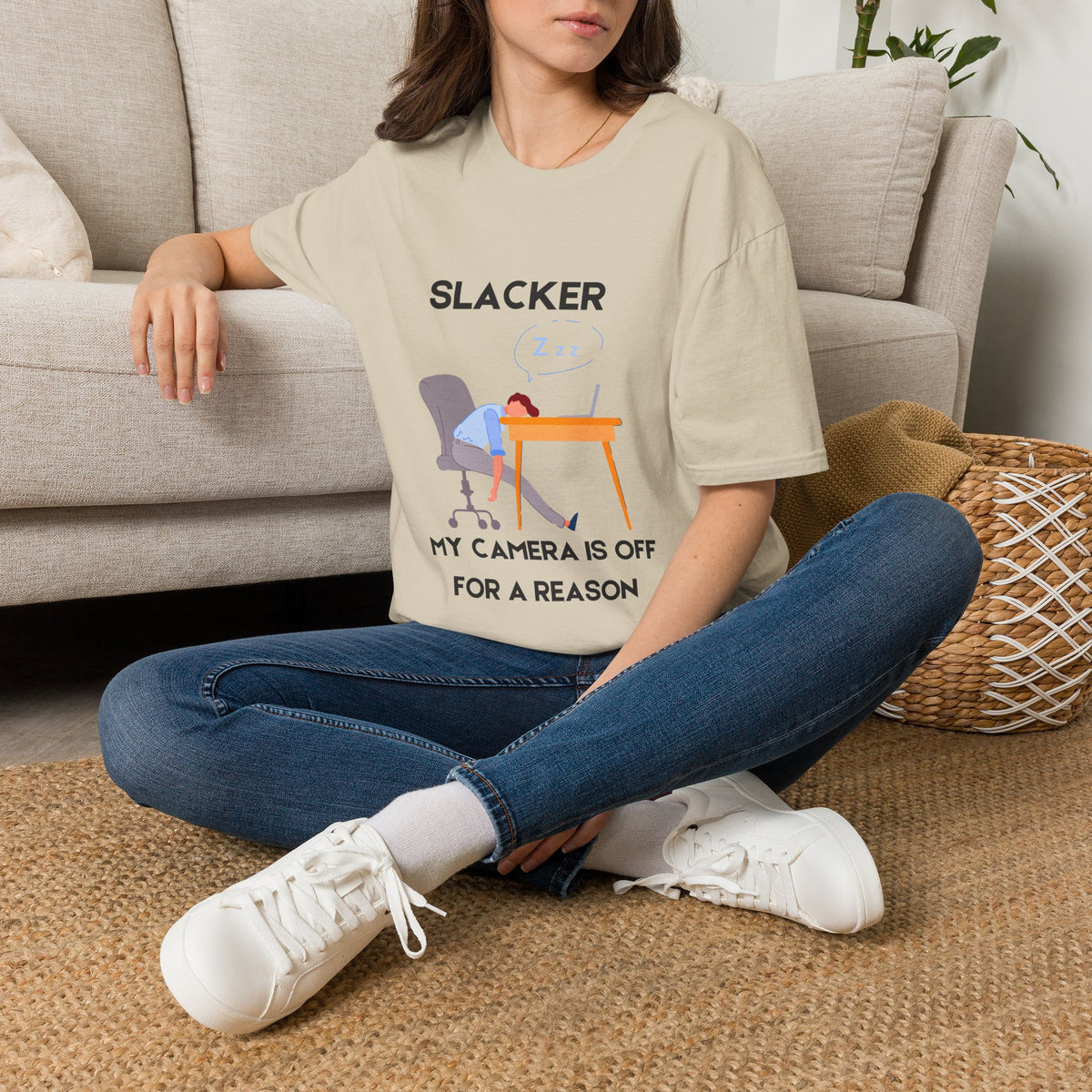 A sand graphic t-shirt displayed on a gray wood plank background, featuring a cartoon illustration of a person sleeping at a desk with the text 'SLACKER, MY CAMERA IS OFF FOR A REASON'. 