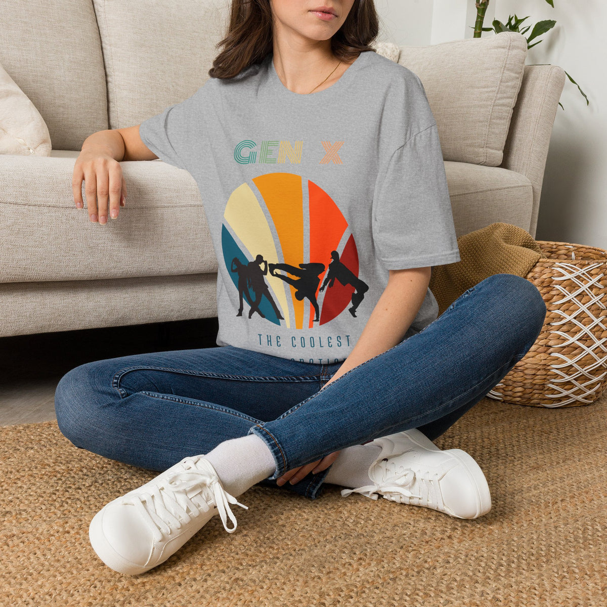 A woman sitting cross-legged on a woven rug in front of a beige sofa, wearing a gray graphic T-shirt with a retro sunset design and the text 'GEN X THE COOLEST GENERATION.