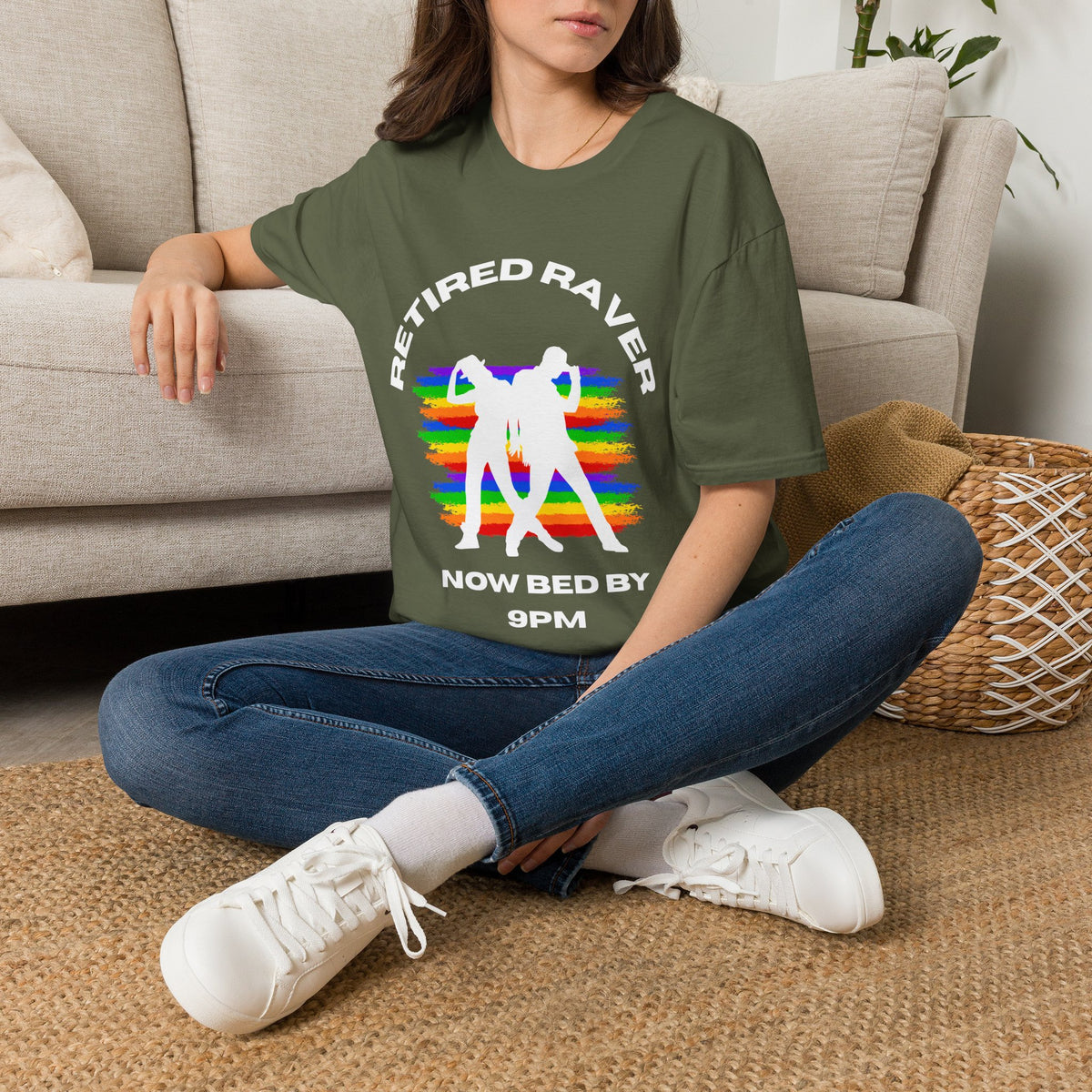 A woman sitting cross-legged on a woven rug in front of a beige sofa, wearing a green graphic T-shirt with white text reading 'RETIRED RAVER NOW BED BY 9PM' and a rainbow-striped dancing figure design.
