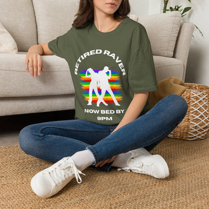 A woman sitting cross-legged on a woven rug in front of a beige sofa, wearing a green graphic T-shirt with white text reading 'RETIRED RAVER NOW BED BY 9PM' and a rainbow-striped dancing figure design.
