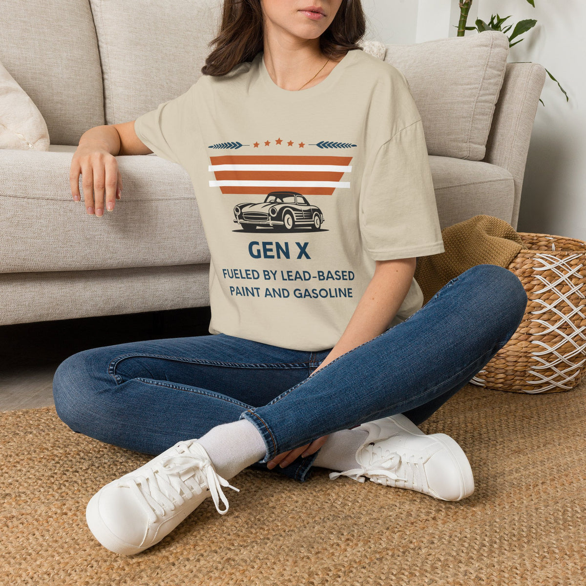 A woman sat on a rug next to a couch wearing a sand graphic t-shirt. The t-shirt features a retro design with the text 'GEN X FUELED BY LEAD-BASED PAINT AND GASOLINE' and an illustration of a classic car under orange and white stripes.