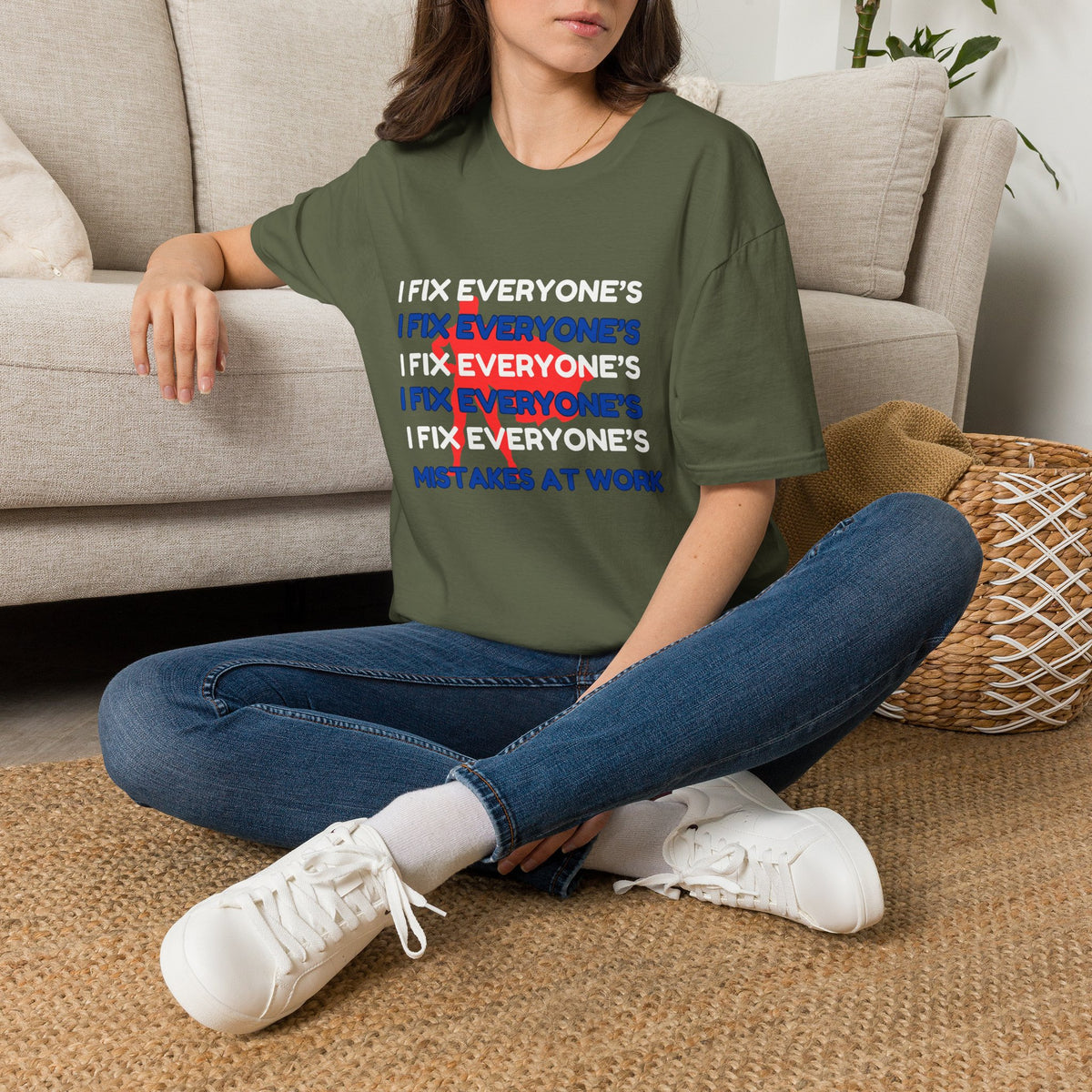 A woman sitting cross-legged on a woven rug in front of a beige sofa, wearing an olive green graphic T-shirt with stacked white, red, and blue text reading 'I FIX EVERYONE'S MISTAKES AT WORK,' along with blue jeans and white sneakers.
