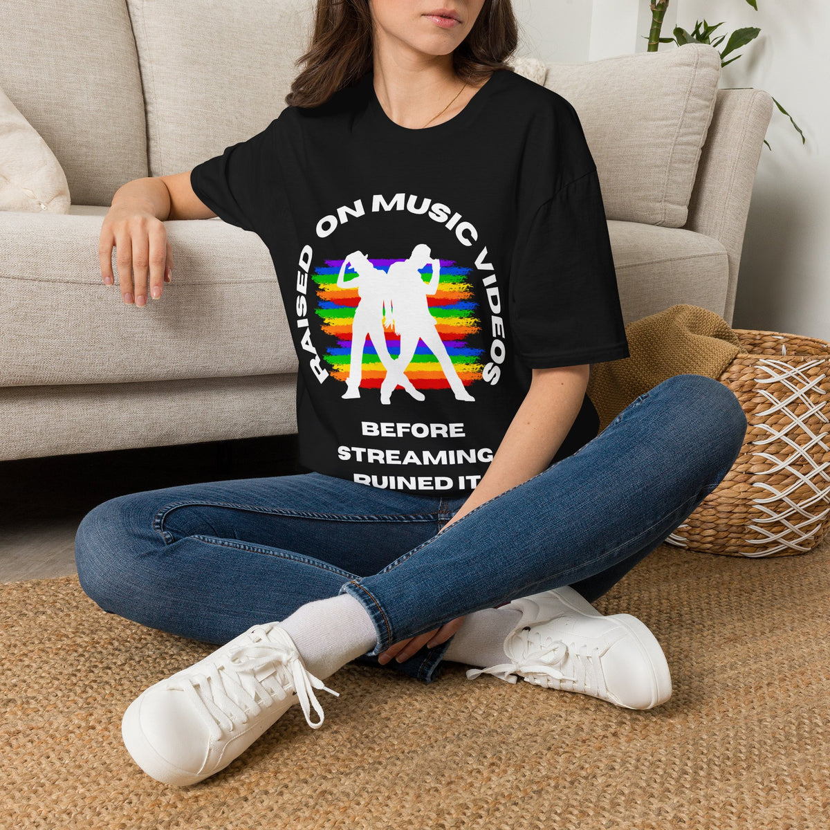 A woman sitting cross-legged on a woven rug in front of a beige sofa, wearing a black graphic T-shirt with white text and a rainbow-striped silhouette design, paired with blue jeans and white sneakers.