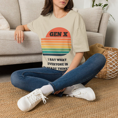 A woman sitting cross-legged on a woven rug in front of a light gray sofa, wearing an oversized sand graphic T-shirt with a retro sunset design and the text 'GEN X I SAY WHAT EVERYONE IS ALREADY THINKING'.