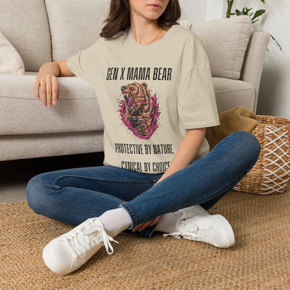 A woman sitting cross-legged on a woven rug in front of a light beige sofa, wearing a beige graphic T-shirt with the text 'GEN X MAMA BEAR' and 'PROTECTIVE BY NATURE. CYGNICAL BY CHOICE.
