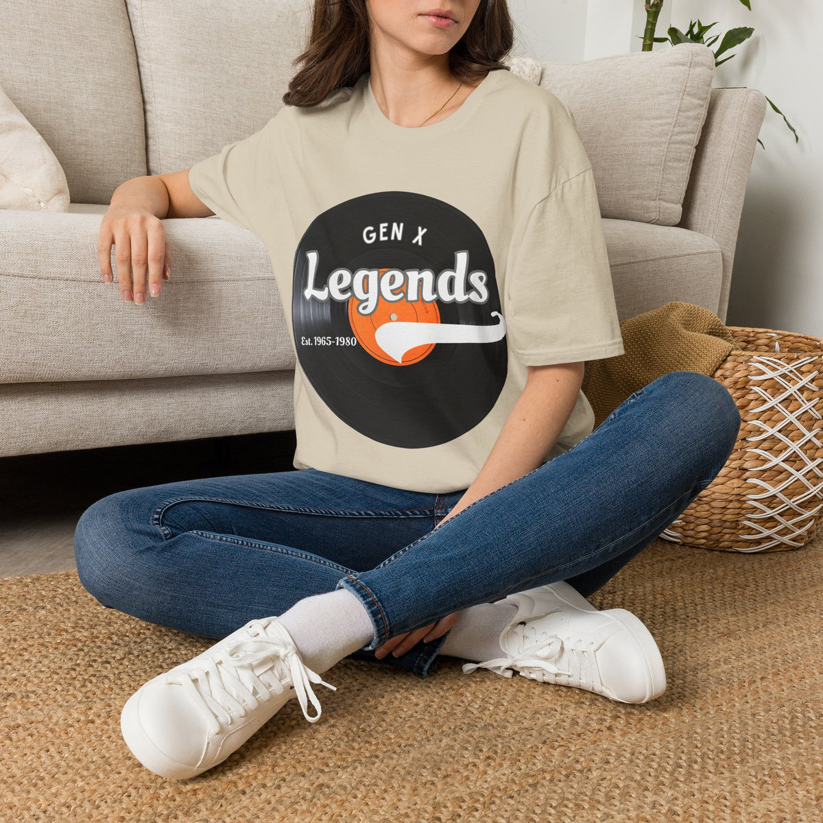 A woman sitting cross-legged on a woven rug in front of a beige sofa, wearing an oversized light sand graphic T-shirt with a vinyl record reading 'GEN X Legends est 1965-1980' 