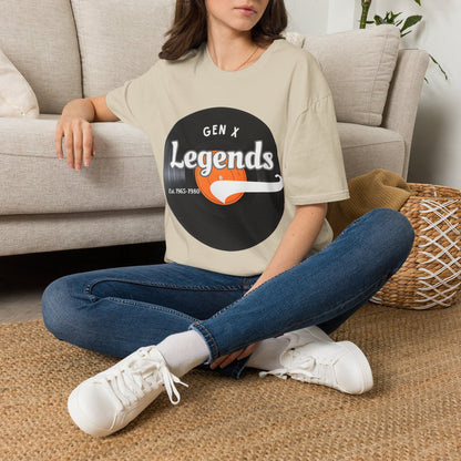 A woman sitting cross-legged on a woven rug in front of a beige sofa, wearing an oversized light sand graphic T-shirt with a vinyl record reading 'GEN X Legends est 1965-1980' 