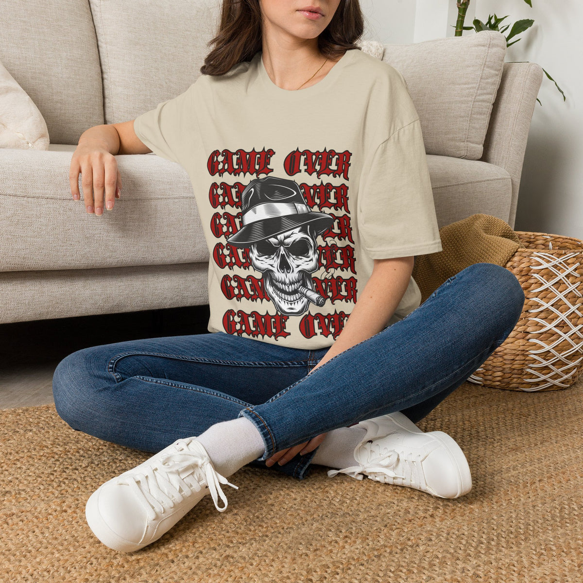 A woman sitting cross-legged on a woven rug in front of a beige sofa, wearing a sand graphic T-shirt with a repeating red 'GAME OVER' design and a black-and-white skull wearing a hat. She is also wearing blue jeans and white sneakers.
