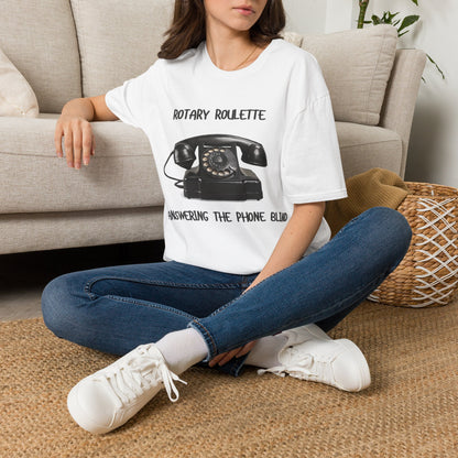 A woman sat on a rug next to couch wearing a white graphic t-shirt featuring a black rotary telephone and the text 'ROTARY ROULETTE' and 'ANSWERING THE PHONE BLIND'.