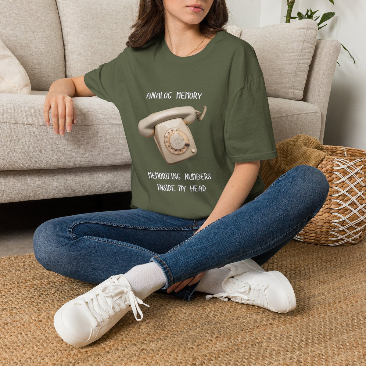 Person wearing a green t-shirt with a green short-sleeve graphic T-shirt The shirt has a black printed design featuring a vintage rotary telephone and the text 'ANALOGIC MEMORY' and 'MEMORIZING NUMBERS INSIDE MY HEAD graphic design sitting on the floor.