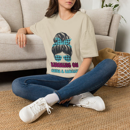 A beige short-sleeve graphic t-shirt  featuring a cartoon illustration of a woman with a plaid hair bow and sunglasses, with the text 'RUNNING ON COFFEE & SARCASM'. 